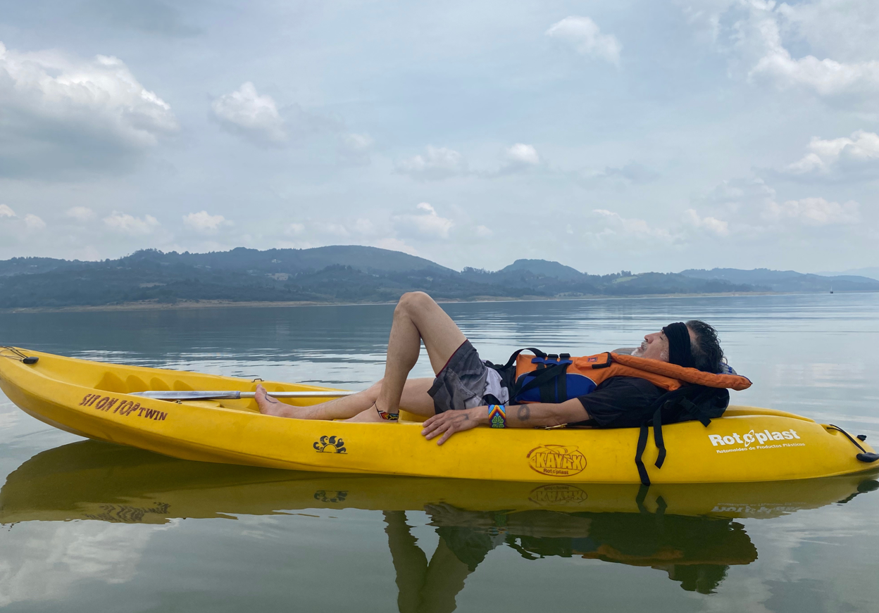 Un hombre recostado de espaldas y totalmente relajado sobre un kayak en medio de un lago tranquilo. Tiene los ojos cerrados y los brazos extendidos, rodeado por el reflejo de las nubes en el agua y un paisaje montañoso al fondo.